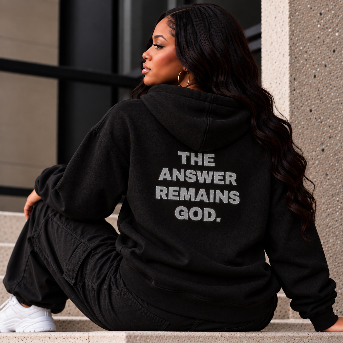 Woman wearing a black hoodie with “THE ANSWER REMAINS GOD.” in silver glitter lettering on the back.