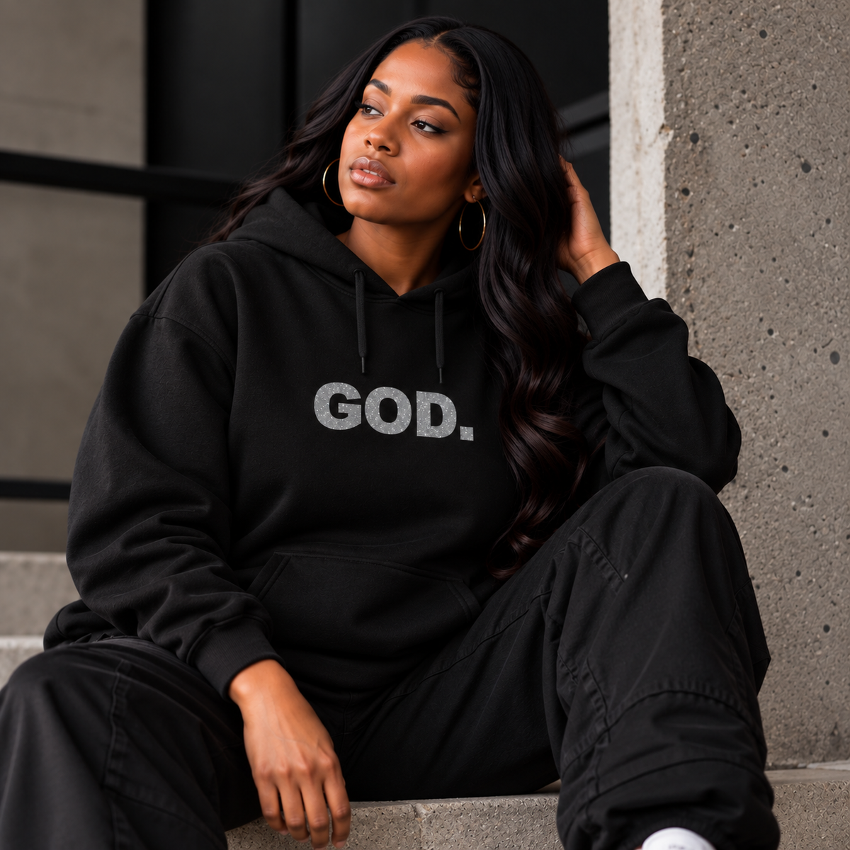 Woman seated on steps wearing a black The Answer Remains hoodie with “GOD.” in silver glitter lettering across the chest.